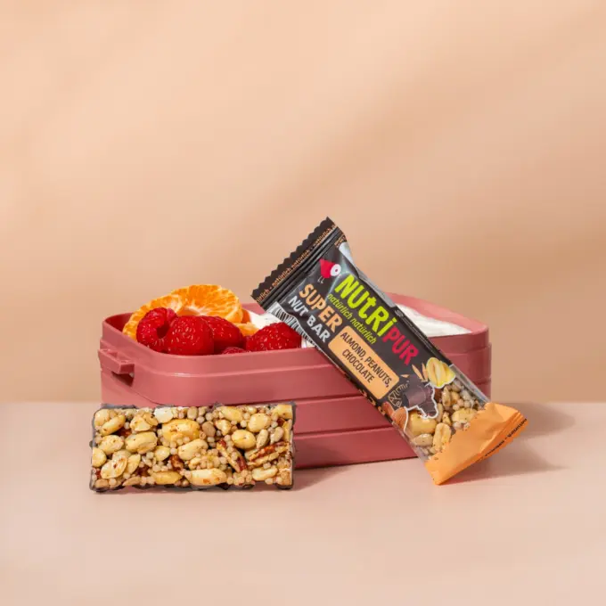 Billedet viser Super Nut nøddebarren med mandler, jordnødder og chokolade med emballage og madkasse.